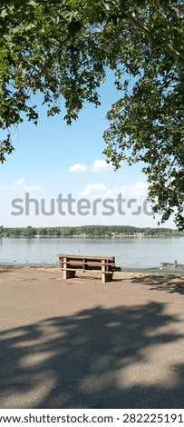 A peaceful riverside landscape featuring an empty wooden park bench facing a wide calm river under a blue sky with light clouds. Green tree branches frame the top of the image, creating a serene and relaxing summer atmosphere.