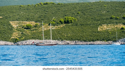 A peaceful sailing boat anchored near a lush green coastline with hills and blue waters. - Powered by Shutterstock