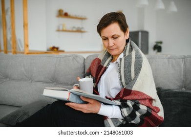Peaceful satisfied mature woman wrapped warm blanket reading book, favorite literature and drinking tea or coffee, happy middle aged senior female relaxing, enjoying free time, weekend at home