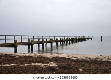 A peaceful seaside scene featuring a weathered wooden pier extending into still ocean waters, under an overcast sky. The tranquil setting conveys a sense of solitude and relaxation. - Powered by Shutterstock