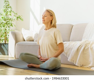 Peaceful senior woman in lotus position meditation with closed eyes at home while sitting on yoga mat on floor, full length. Calm elderly lady practicing meditation techniques and yoga indoors - Shutterstock ID 1984633001