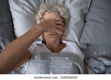Peaceful sleepy senior woman awaking in her bed, rubbing face, covering eyes with hand from disturbing light. Elderly mature lady suffering from insomnia, sleep disorder or deprivation. Top view