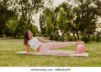 peaceful, smiling woman in pink clothes and small rubber ball doing pilates on pink mat at sunset in park in summer. 