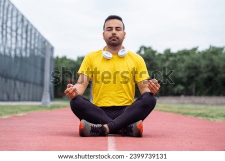 Peaceful sportsman, donning athletic attire, practices mindfulness in lotus pose on a green park's running track.