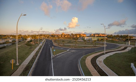 A peaceful suburban area during sunset, featuring a newly paved road with modern streetlights, surrounded by landscaped greenery and palm trees. Houses in the distance are bathed in golden sunlight. - Powered by Shutterstock