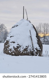 A peaceful vertical shot of a large haystack partially covered with fresh white snow at dawn. The background features a beautiful winter sunrise with golden light on the horizon. An authentic scene of a cold morning in the countryside, ideal for agriculture, nature, and winter holiday themes.