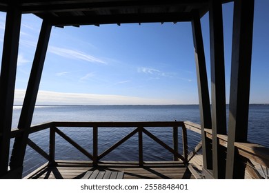 A peaceful view from a wooden dock, looking out over the calm water with a clear blue sky and a gentle reflection on the surface. The dock’s railing frames the tranquil scene. - Powered by Shutterstock