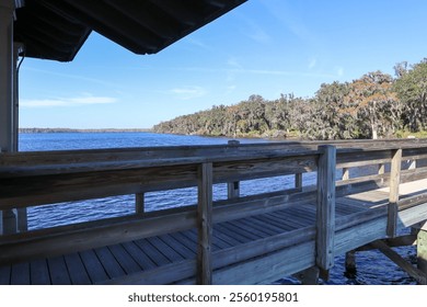 A peaceful view from a wooden dock, overlooking calm river waters with trees lining the shore under a bright blue sky. The wooden railing adds a rustic touch to the scenic landscape. - Powered by Shutterstock