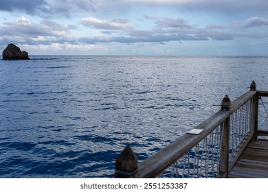 A peaceful view from a wooden pier overlooking a vast ocean at dusk. The water reflects soft light as clouds drift across the sky, highlighting a distant rock formation in the sea. - Powered by Shutterstock