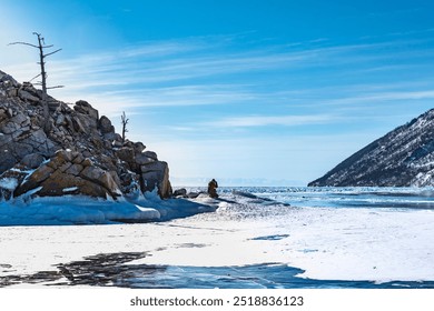 A peaceful winter scene with a lone person sitting on rocky terrain near a frozen lake. The barren trees and icy shoreline contrast against the vast blue sky and distant snow-covered mountains. - Powered by Shutterstock