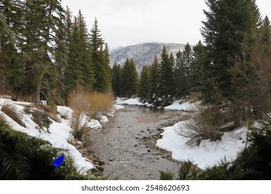 Peaceful winter scene with a snow-covered creek surrounded by evergreen trees and snowy mountains under a bright blue sky with soft clouds. - Powered by Shutterstock