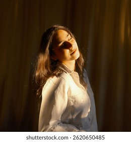 Mujer tranquila en camisa blanca disfrutando de la luz dorada del sol con los ojos cerrados y la expresión serena, de pie contra el telón de fondo de la cortina en luz interior cálida suave
