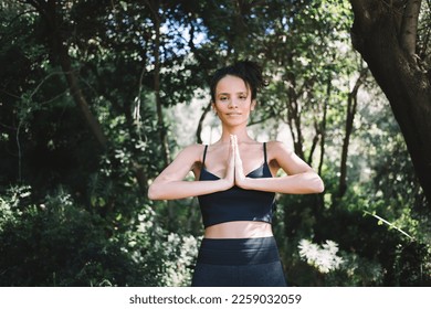Peaceful young ethnic female in activewear standing with hands clasped and practicing namaste gesture while looking at camera in forest - Shutterstock ID 2259032059