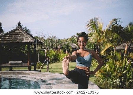 Peaceful young ethnic female in activewear doing Standing Hand to Big Toe pose while practicing yoga on poolside against tropical trees