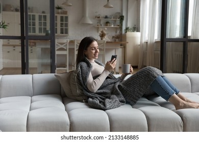 Peaceful young female relaxing on cozy sofa covered with knitted warm plaid, holding smart phone and teacup drinking tea spend carefree free time alone at home. Weekend leisure, modern tech concept - Shutterstock ID 2133148885