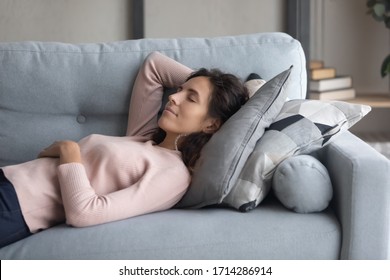 Peaceful young happy woman lying on soft pillow on comfortable sofa in living room, enjoying lazy weekend time. Calm pleasant millennial girl napping daydreaming alone on couch, resting indoors.