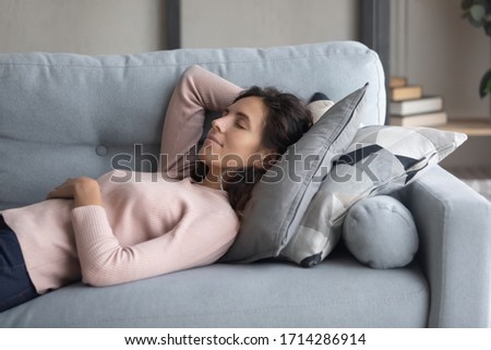 Peaceful young happy woman lying on soft pillow on comfortable sofa in living room, enjoying lazy weekend time. Calm pleasant millennial girl napping daydreaming alone on couch, resting indoors.