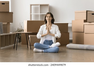 Peaceful young home owner woman meditating during stressful moving into new home, sitting on floor, keeping meditation pose, eyes closed, Namaste hand gestures with stacked boxes behind