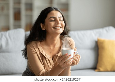 Peaceful young indian lady sitting on couch with closed eyes, enjoy drinking coffee, enjoying morning with warm drink in living room, copy space - Powered by Shutterstock