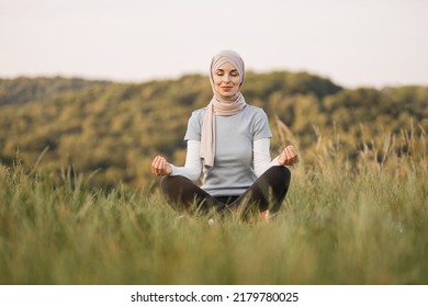 Peaceful Young Lady In Hijab Meditating With Closed Eyes In The Park. Pretty Muslim Woman Sitting On Mat In Yoga Pose, Finding Inner Balance.