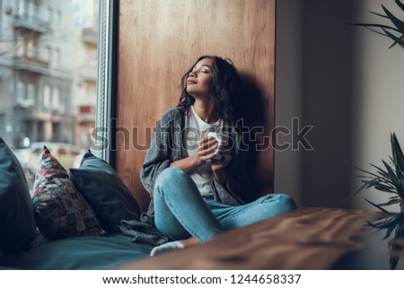 Peaceful young lady sitting on the window sill and smiling while drinking tea with her eyes closed