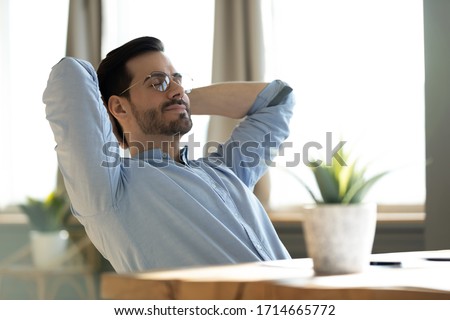 Peaceful young man wearing glasses daydreaming with closed eyes, lazy sleepy businessman or student leaning back in comfortable chair, stretching hands, sitting at work desk, dreaming and visualizing