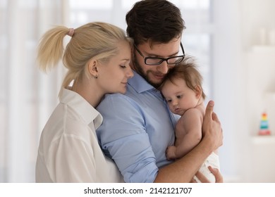 Peaceful young married couple enjoying being family, parents, holding few month baby in arms, standing close together, hugging with closed eyes. Parenthood, childbirth, relationship concept