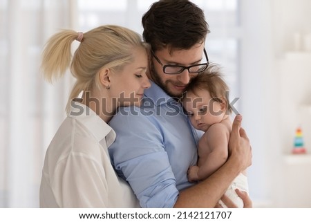 Peaceful young married couple enjoying being family, parents, holding few month baby in arms, standing close together, hugging with closed eyes. Parenthood, childbirth, relationship concept