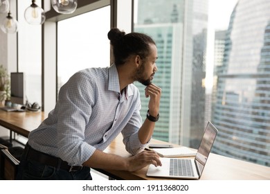 Peaceful young mixed race guy stand by desk distracted from pc admire stunning urban city view from home office. Pensive young black man rest from computer work looking through panoramic window wall