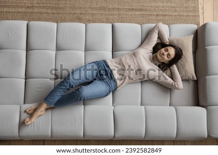 Peaceful young woman in casual clothes relaxing alone on cozy sofa with hands behind head and eyes closed, above view. Fatigue relief, comfortable furniture store ad, carefree leisure at home concept