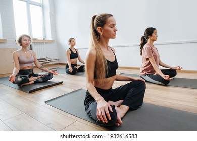 Peaceful young women practicing yoga in a group of four, sitting on a floor with legs closely tucked. Meditation, relaxing, clearing consciousness. Eyes closed, hands on knees.