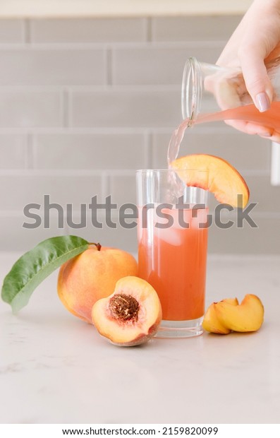 A peach\
drink being poured into a glass of ice. Peaches spread around the\
countertop. A human hand doing the\
pouring
