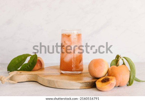 A Peach drink in a glass with ice in a\
marble kitchen. There are also fresh fruit, some cut up. The drink\
is on a brown cutting board, ready to serve.\

