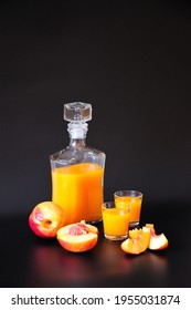 Peach liqueur in a transparent glass bottle and two glasses, next to it are pieces of ripe fruit on a black background. Close-up.