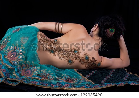 A peacock feather henna design on a woman's back. She is wearing a teal colored sari. Shot in the studio on a black background.