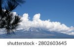 Snow‑capped peak beneath bright clouds — A towering mountain with streaks of snow rises into thick white clouds under a vivid blue sky, framed by a pine branch and a red fence in the foreground.