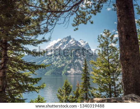 A peak of the Grand Teton mountain range, set upon the stills waters of Phelps Lake, Grand Teton National Park, Wyoming, USA. The snow dappled mountain is framed by the green pine forests.