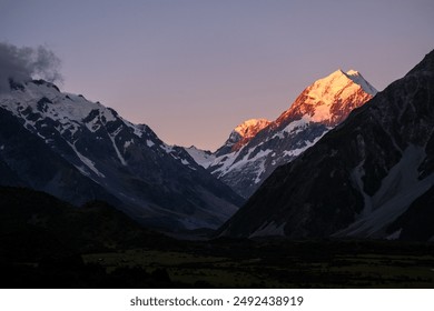 The peaks of Mount Cook are illuminated by the warm glow of sunset, with snow-capped mountains contrasting against a pastel sky. The alpine landscape is serene and majestic. - Powered by Shutterstock