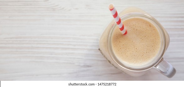 Peanut Butter Banana Smoothie In A Glass Jar On A White Wooden Surface, Top View. Overhead, Flat Lay, From Above. Space For Text.