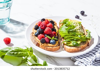 Peanut Butter And Berries Toast And Avocado Toast With Basil On A White Plate. Healthy Vegan Breakfast Concept.