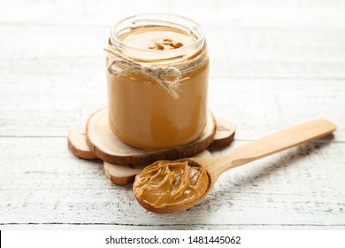 Peanut Butter In Glass Jar With Spoon On White Wooden Table