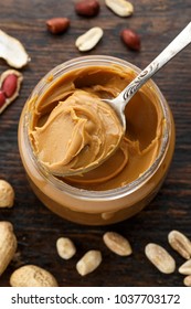 peanut butter in an open jar on a brown wooden background, next to peanuts scattered