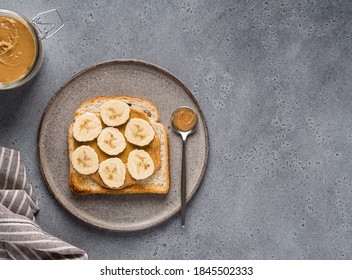 Peanut butter toast with banana slices on ceramic plate with peanut butter jar and tea spoon. Healthy  breakfast or snack. Top view. Copy space. Grey concrete background.