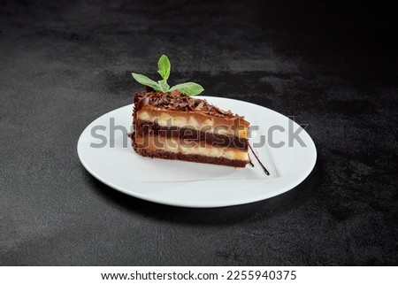 Peanut and caramel cake with chocolate topped on white plate on dark stone background. Piece of Snickers cake in minimal style. Toffee, nuts and chocolate cake in confectionery menu