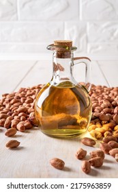 Peanut Oil In A Glass Jug And Raw Peanuts On A White Wooden Table. Monounsaturated Cooking Oil Made Of Arachis Hypogaea. Groundnut As Edible Seeds And Oil Crop. Dietary Nutrition Concept. Front View.