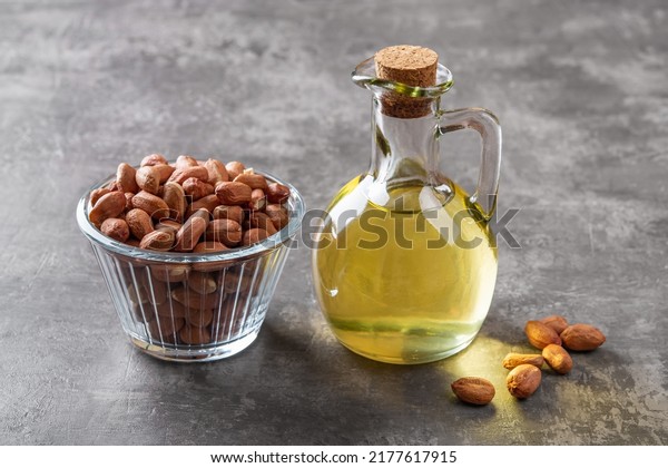 Peanut oil in a glass jug and raw peeled groundnut\
in a glass bowl over gray background. Arachis hypogaea as edible\
seeds and oil crop. Monounsaturated cooking oil. Vegetarian snack.\
Top view.