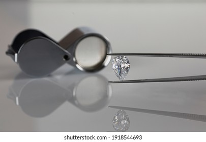 Pear shape polished diamond in tweezers with loupe on the light background. Diamond grading.