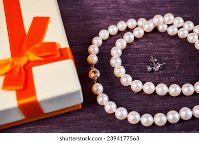Pearl necklace and earrings with small diamonds and a bright gift box with orange bow on wooden table - Shutterstock ID 2394177563
