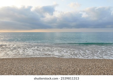 Pebble beach with gentle waves and calm turquoise waters, under a partly cloudy sky, creating a peaceful and serene coastal view - Powered by Shutterstock