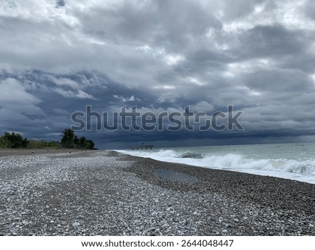 a pebble beach under a stormy, cloudy sky. Waves crash gently onto the shore, reflecting the dark, brooding atmosphere. Hints of distant land and a structure can be seen on the horizon